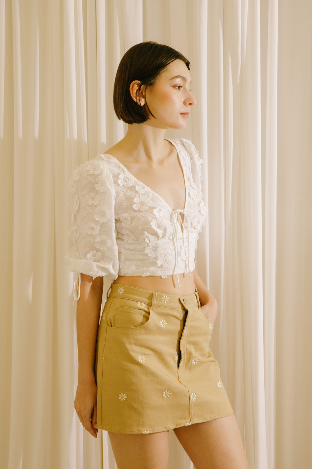 A woman wearing a white crop top with dimensional flower embellishment short-sleeve and a mustard yellow mini skirt with white embroidered details, standing in front of a cream-colored curtain backdrop.