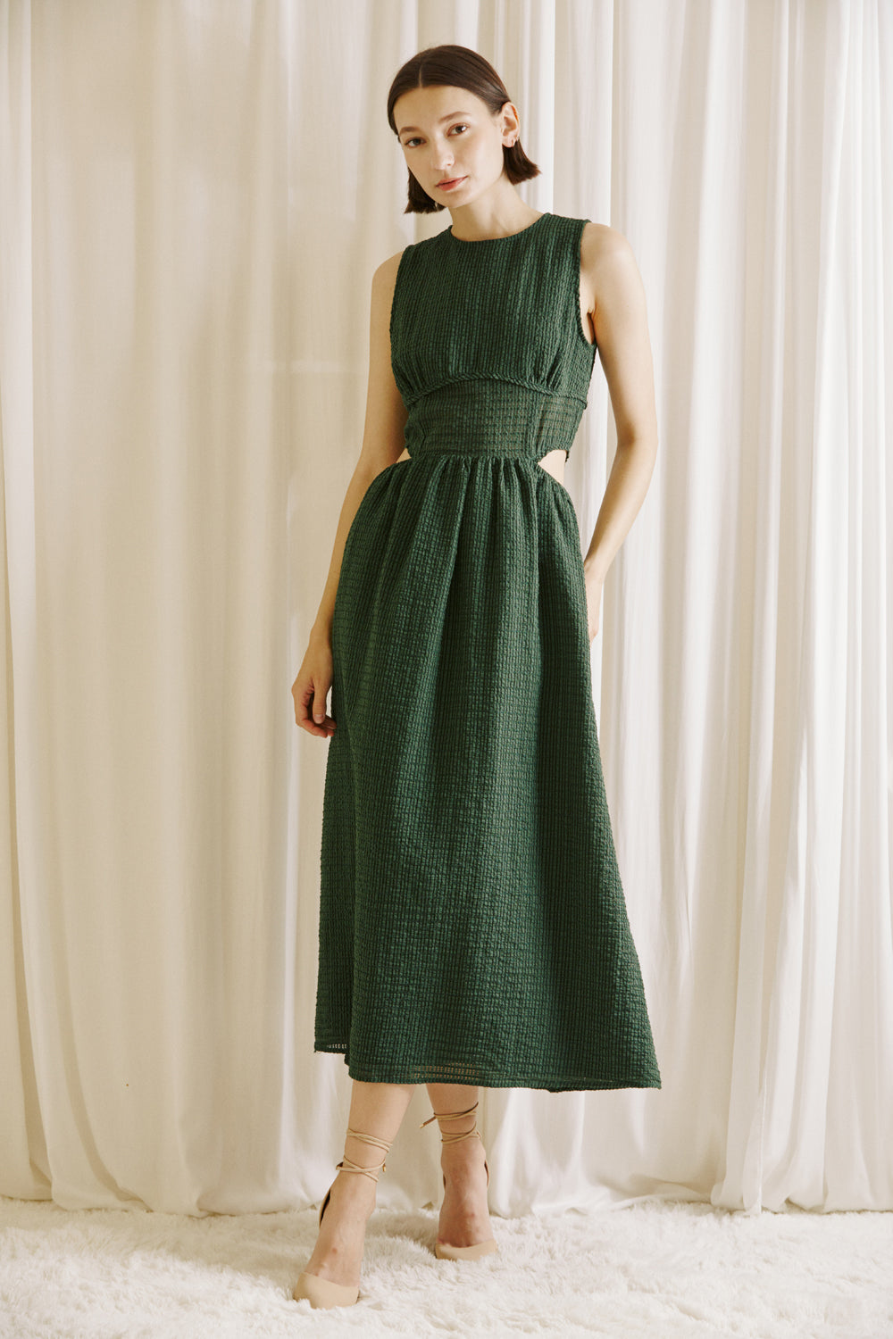Woman wearing a green midi dress with side cut-outs and beige high-heels standing against a white curtain backdrop