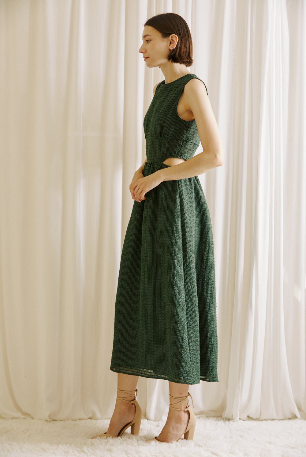 Side view of a woman wearing a green midi dress with side cut-outs and beige high-heels standing against a white curtain backdrop