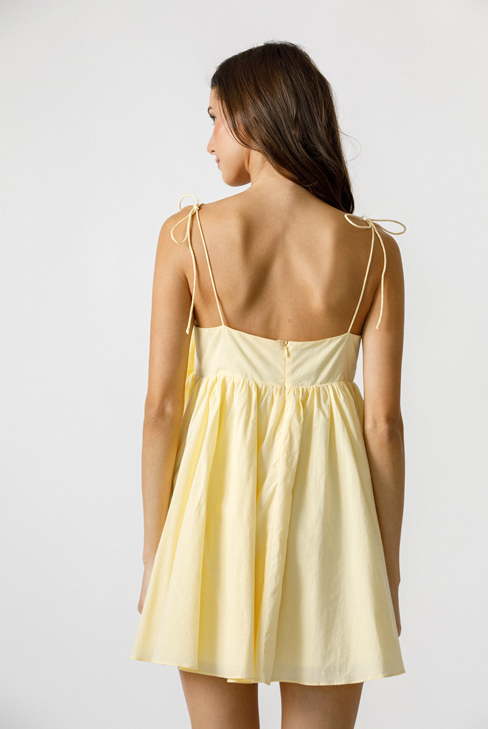 Back view of a woman wearing a light yellow dress with thin straps on a white background