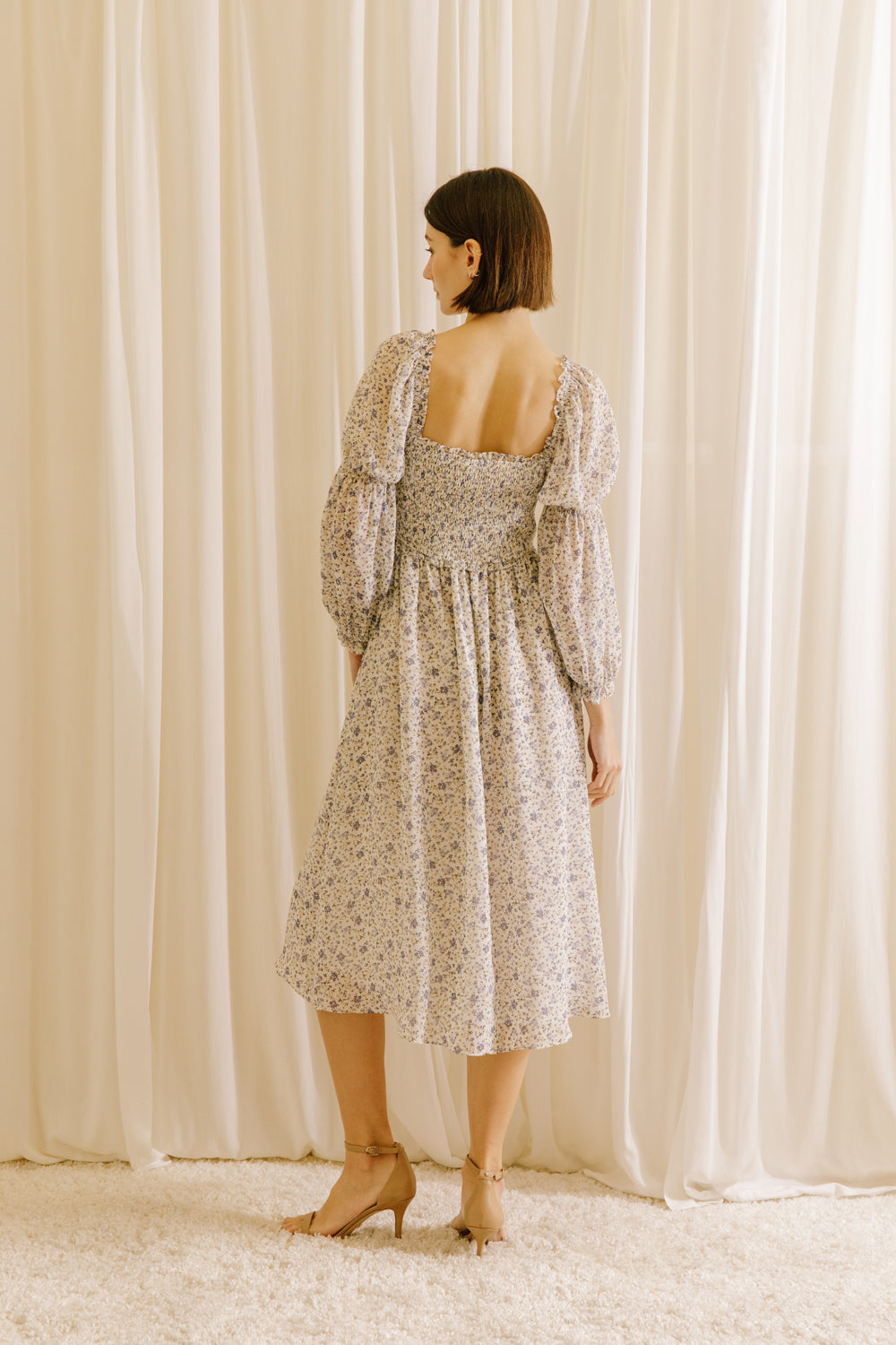 Back view of a woman standing in a room with curtains wearing a beige floral square neckline midi dress with long sleeves.