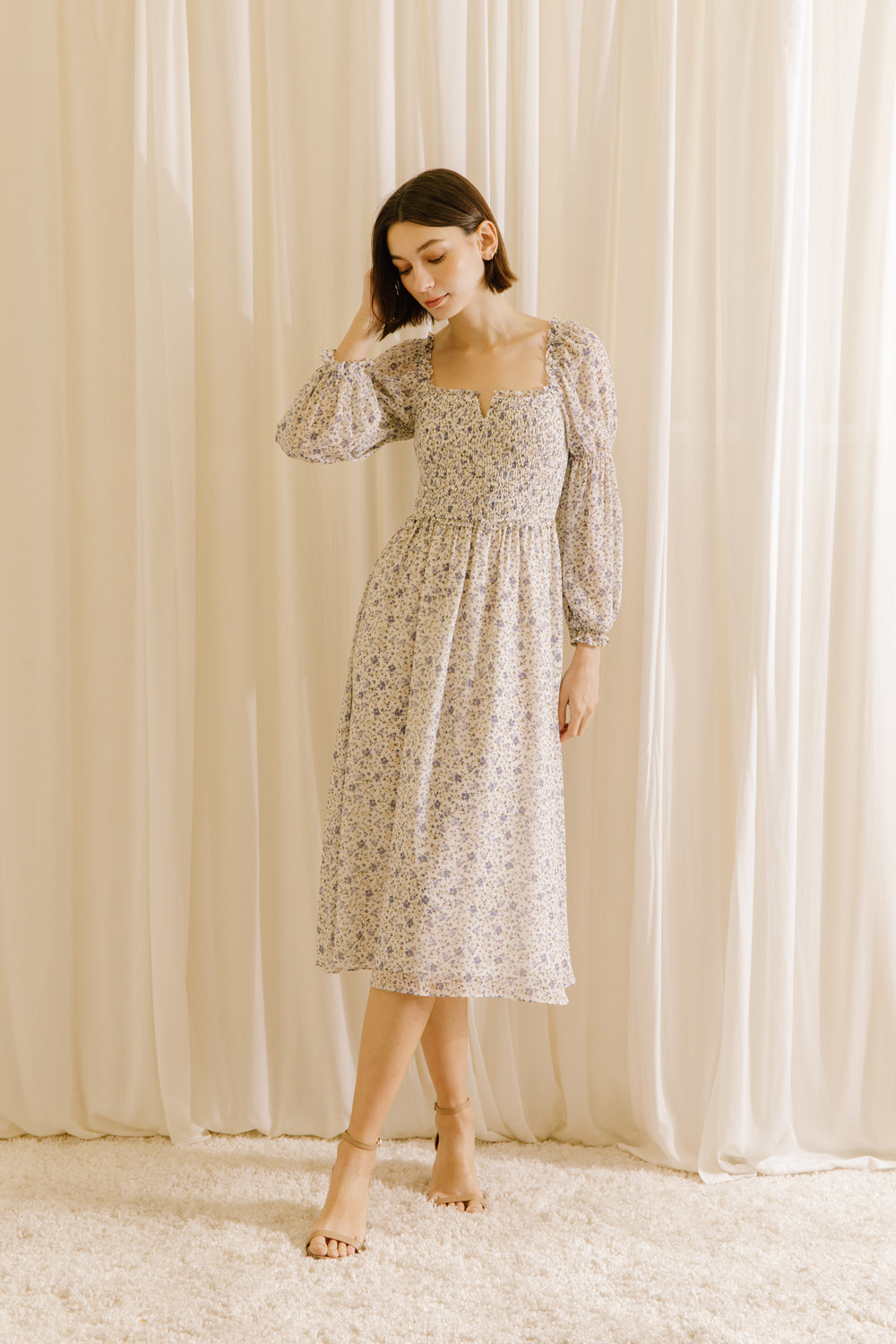 Woman wearing a floral dress with long sleeves standing against a white curtain backdrop