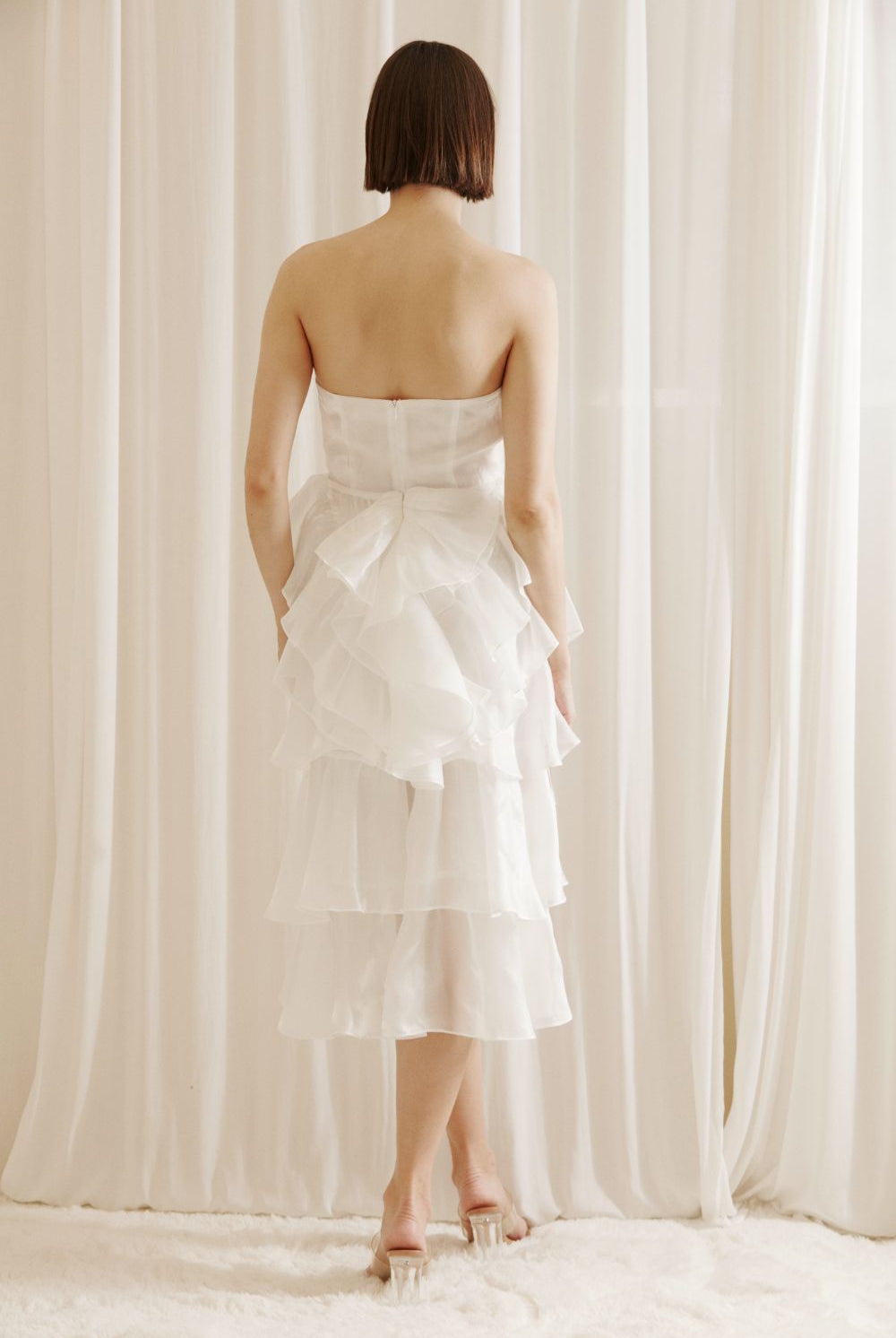 Back view of a woman wearing a white strapless dress with asymmetrical ruffled layers against a white curtain background