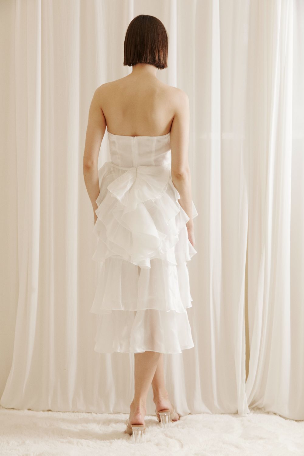 Back view of a woman wearing a white strapless dress with asymmetrical ruffled layers against a white curtain background