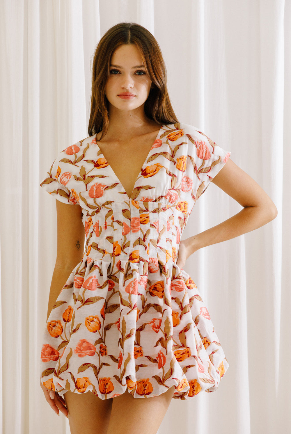 Woman wearing a coral and white floral dress against a white curtain background