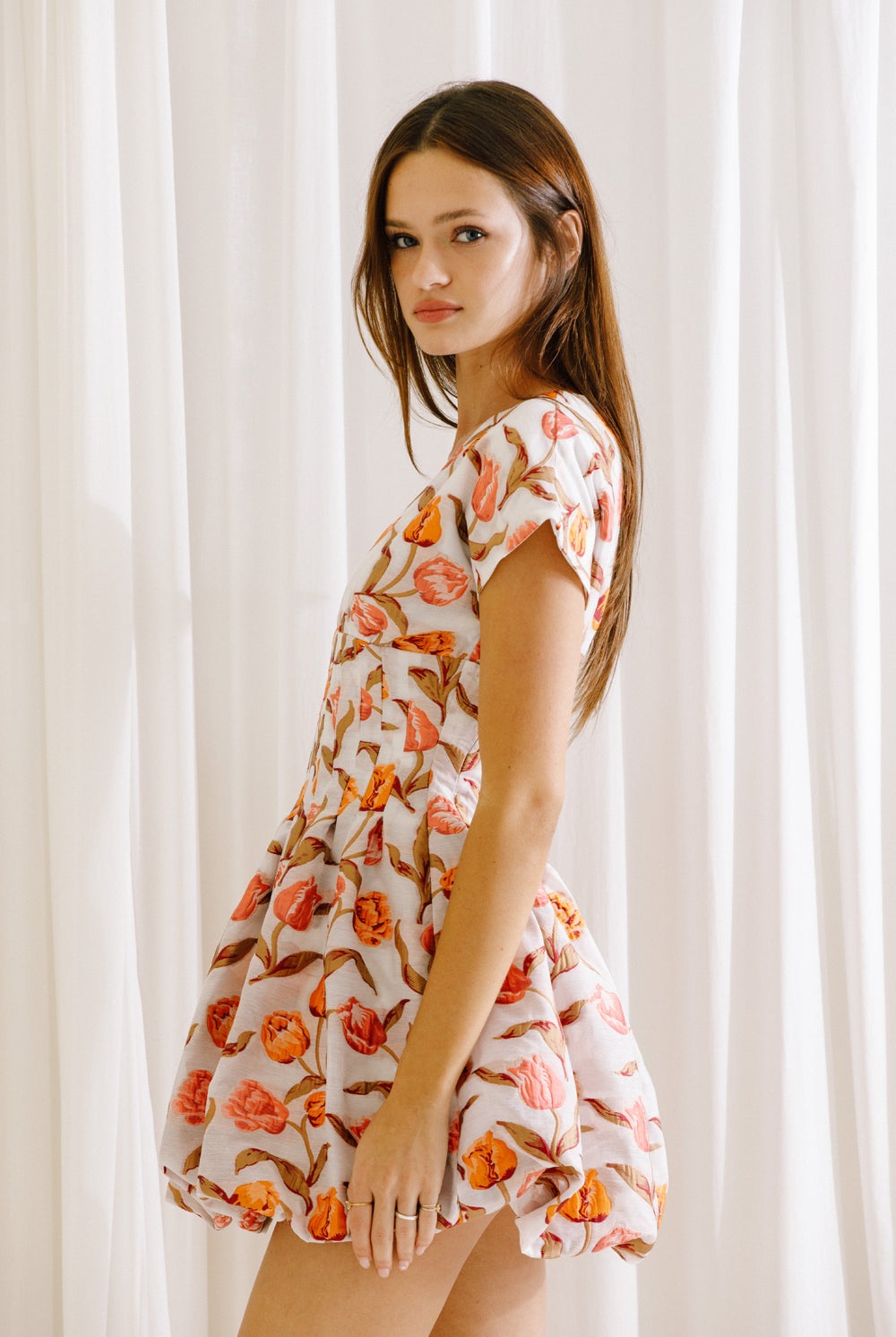 Side view of a woman wearing a coral and white floral dress against a white curtain background