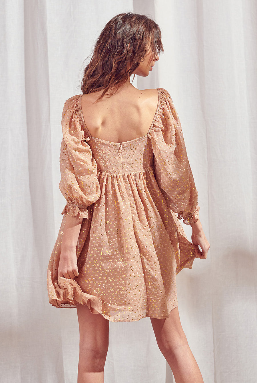 Back view of a woman wearing a brown dress with shimmering gold flaked embroidered dots and an invisible zipper against a white curtain background