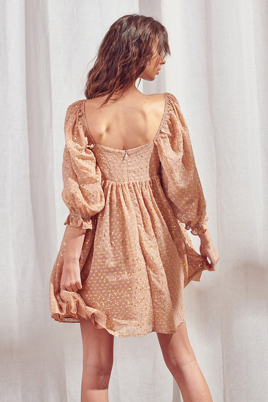 Back view of a woman wearing a brown dress with shimmering gold flaked embroidered dots and an invisible zipper against a white curtain background