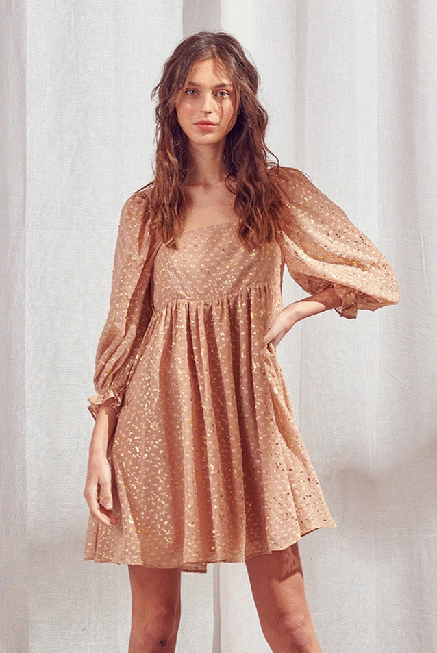 Woman wearing a brown dress with shimmering gold flaked embroidered dots against a white curtain background