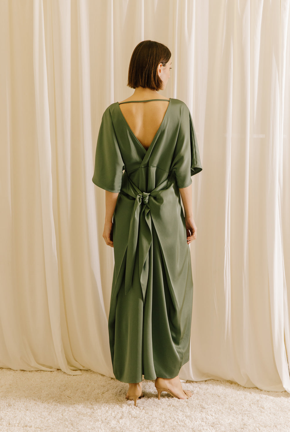 Back view of a woman wearing an olive green V-back maxi dress with short sleeves and fluid drape