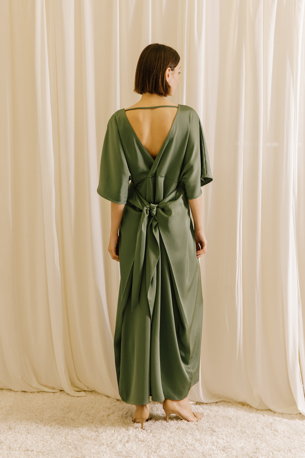 Back view of a woman wearing an olive green V-back maxi dress with short sleeves and fluid drape
