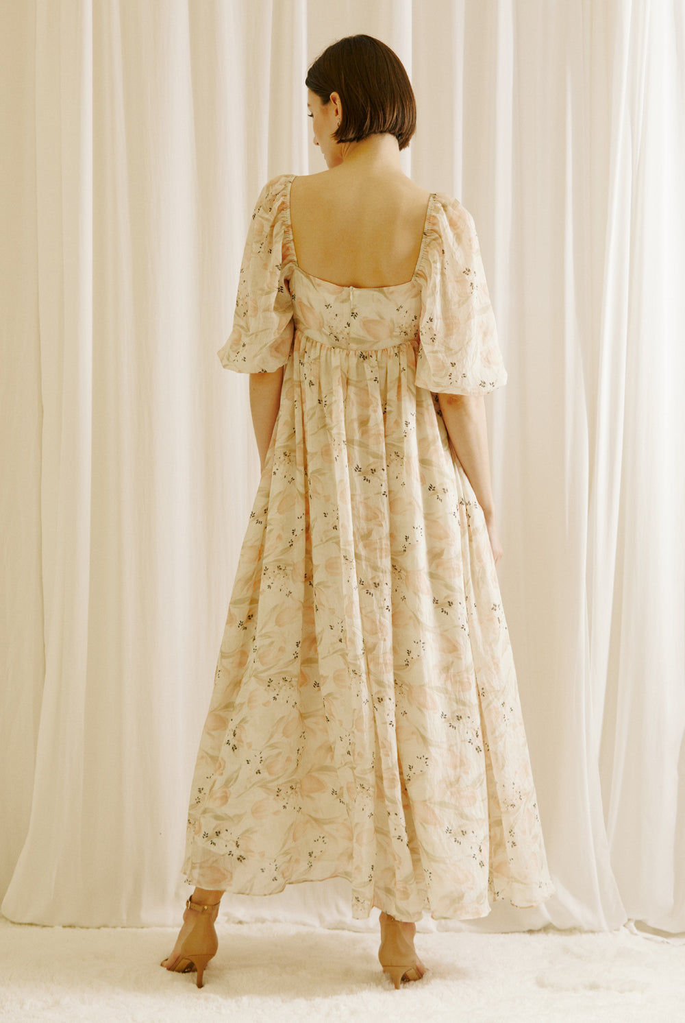 Back view of a woman wearing a floral dress with tulips print standing against a white curtain backdrop