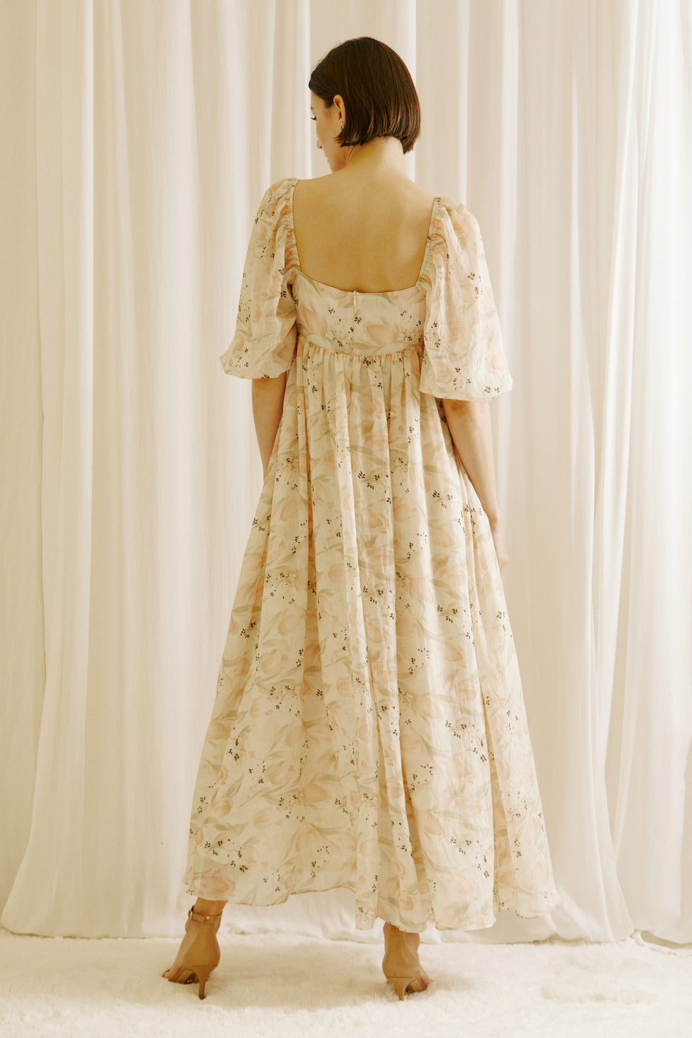 Back view of a woman wearing a floral dress with tulips print standing against a white curtain backdrop