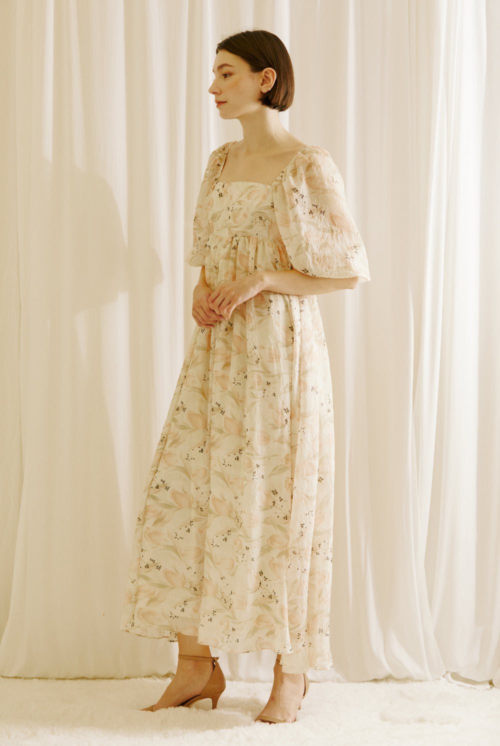 Side view of a model wearing a floral dress with a tulip print standing against a white curtain backdrop