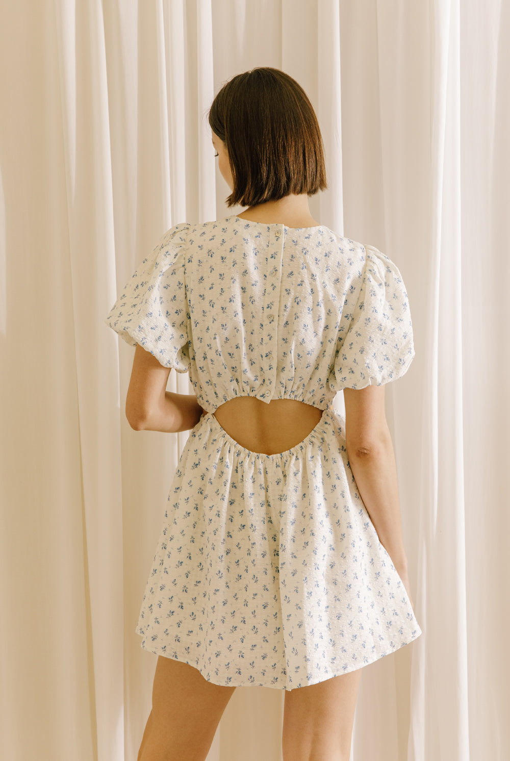 Back view of a model wearing a cream dress with a floral pattern and cut-outs to the waist against a beige curtain background