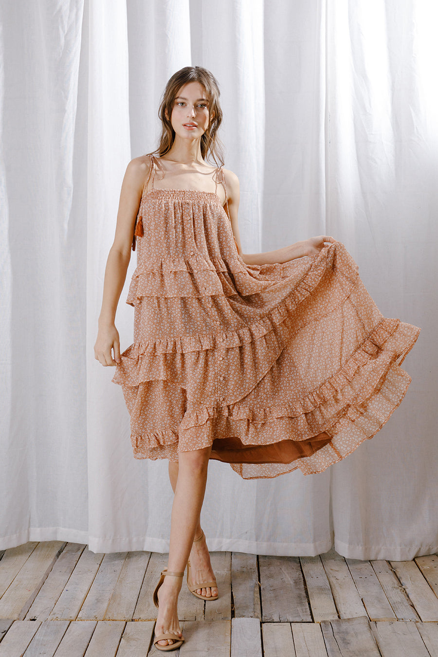 A model wears a dreamy warm-weather midi dress featuring a vintage floral print, square neckline, and flowy ruffled layers against a white curtain.