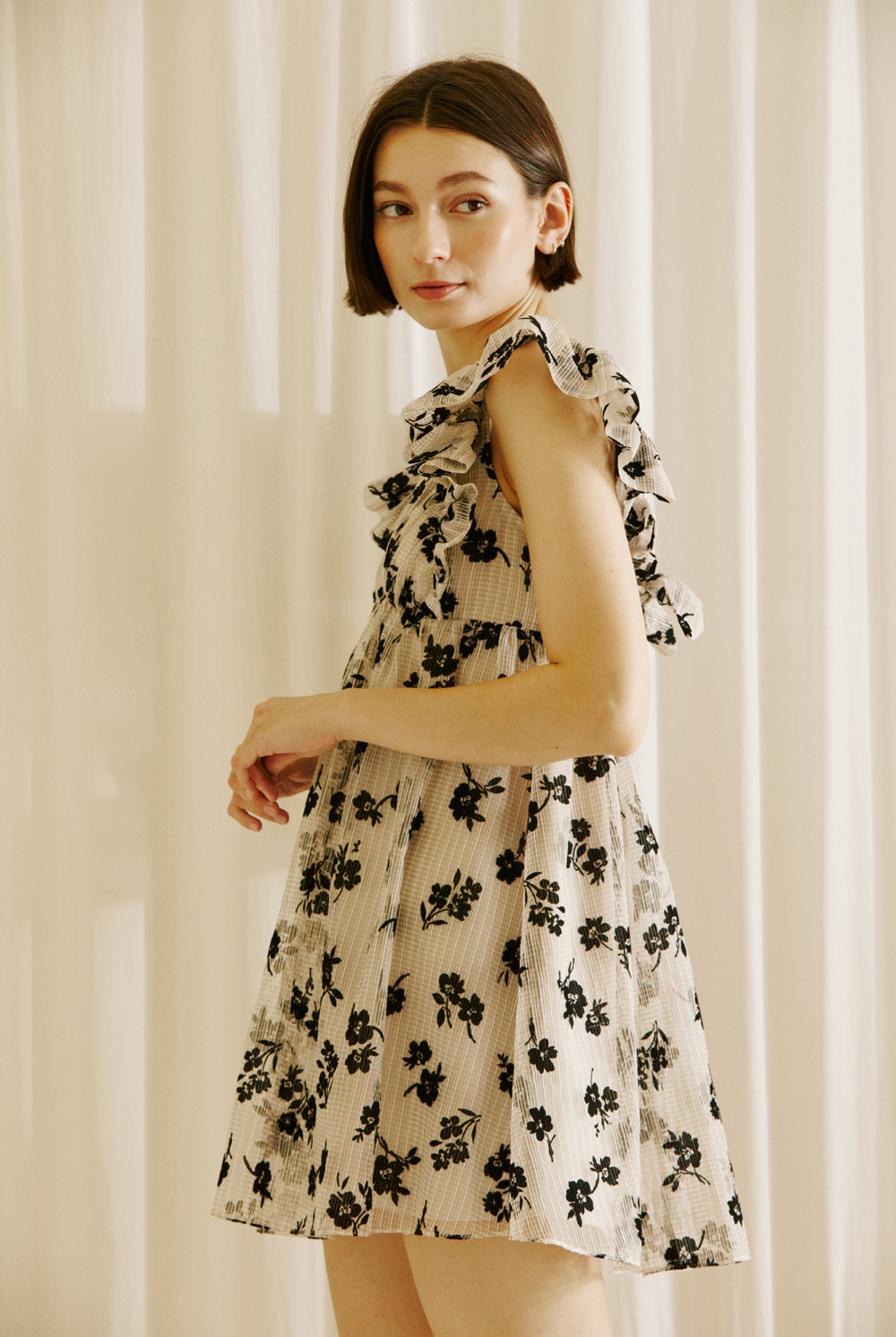 Side view of a woman wearing a beige dress with black floral sheer print, v-neckline and ruffled cap sleeves against a plain background