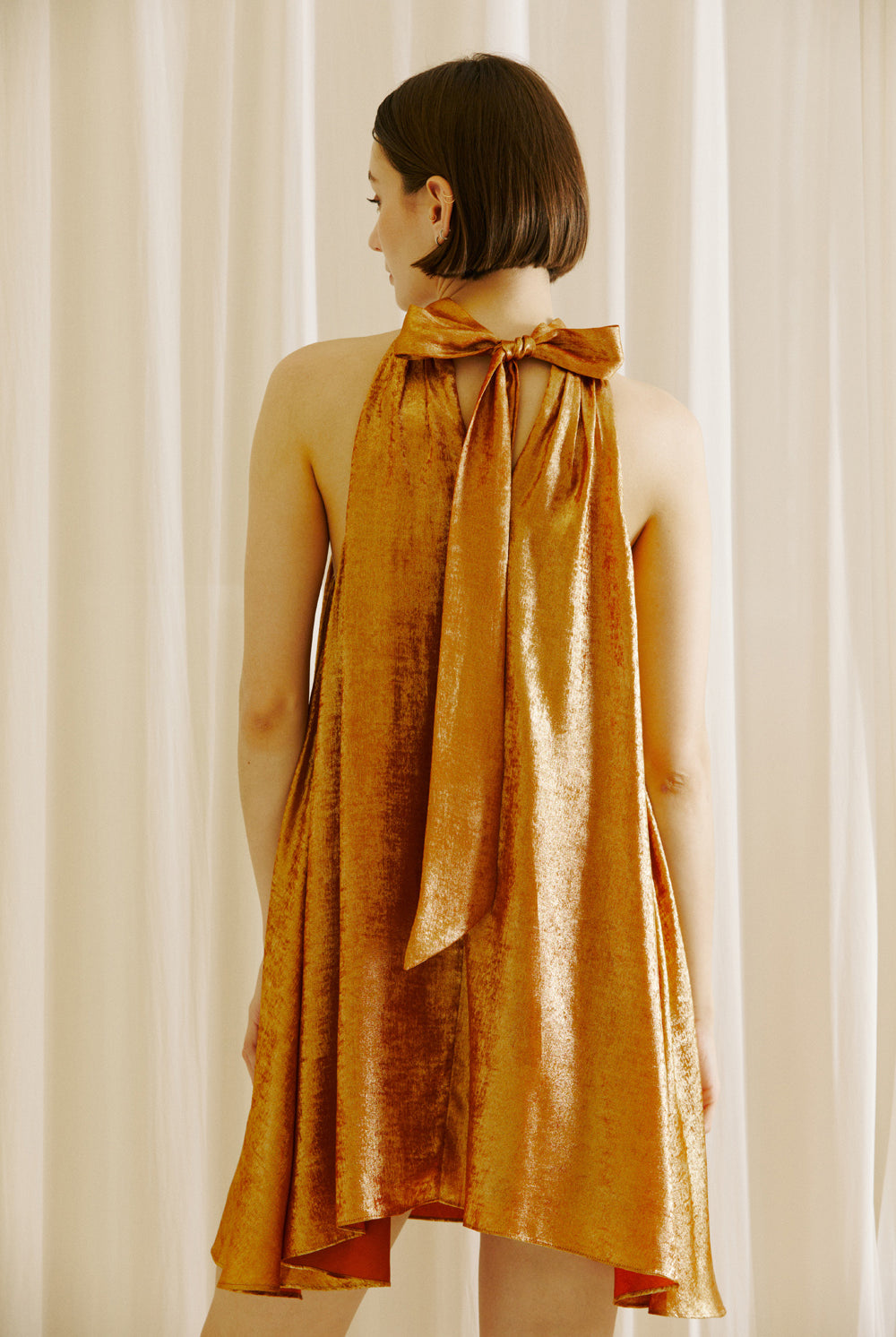 Back view of a woman wearing a bronze halter neck dress against a white curtain background