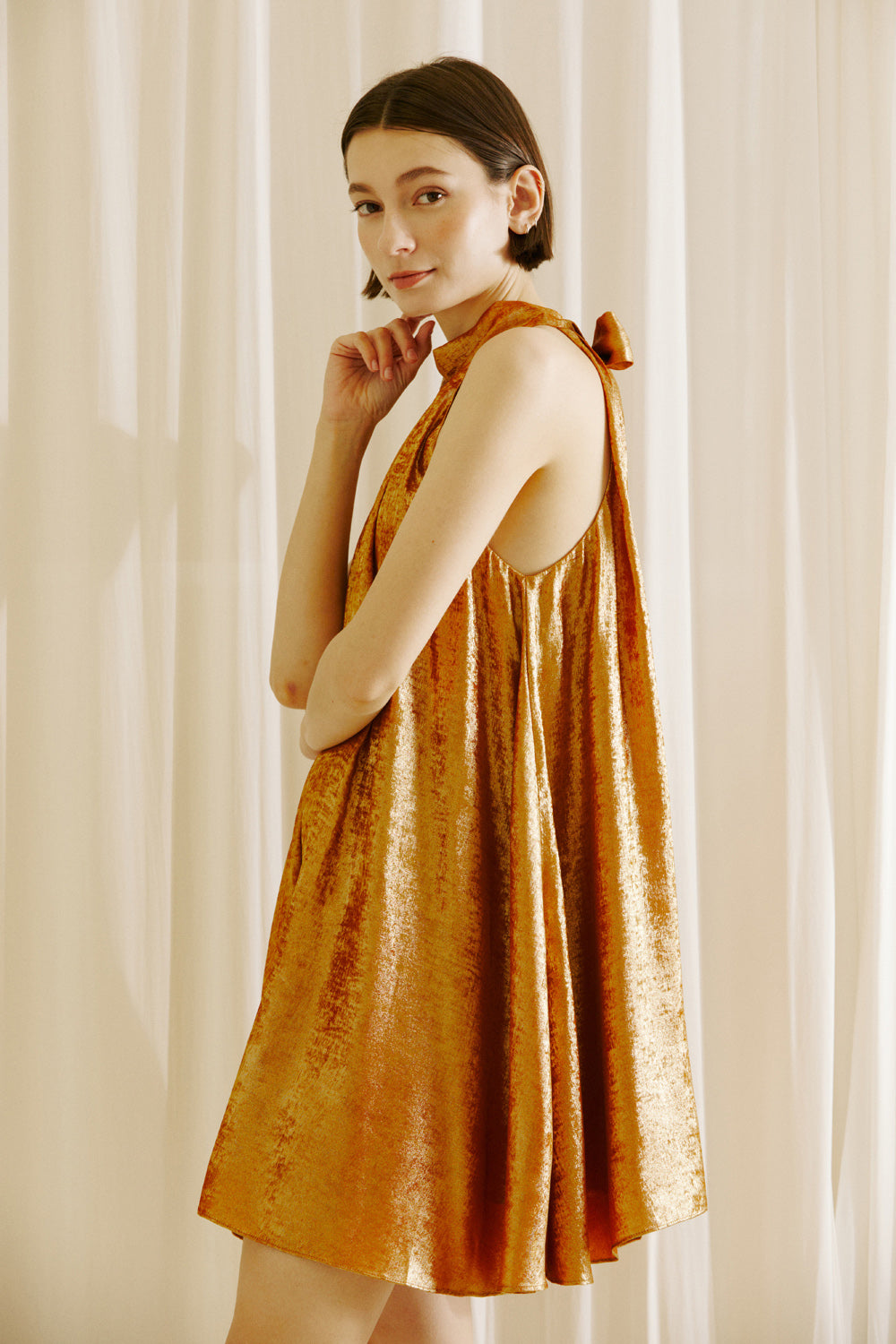 Side view of a woman wearing a bronze halter neck dress against a white curtain background