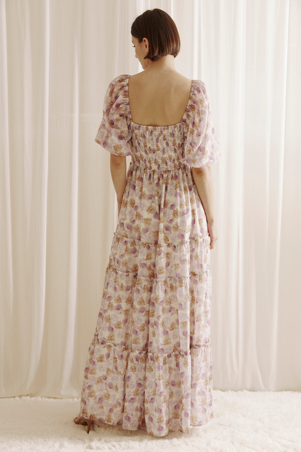 Back view of a woman wearing a purple floral dress with a slit standing against a white curtain.