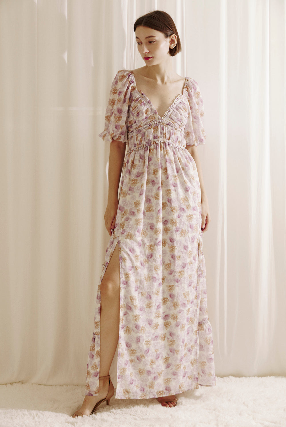Woman wearing a purple floral dress with a slit standing against a white curtain.