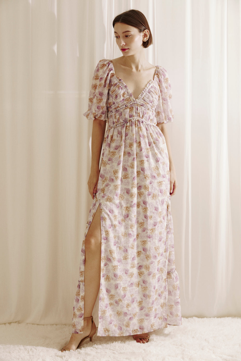 Woman wearing a purple floral dress with a slit standing against a white curtain.