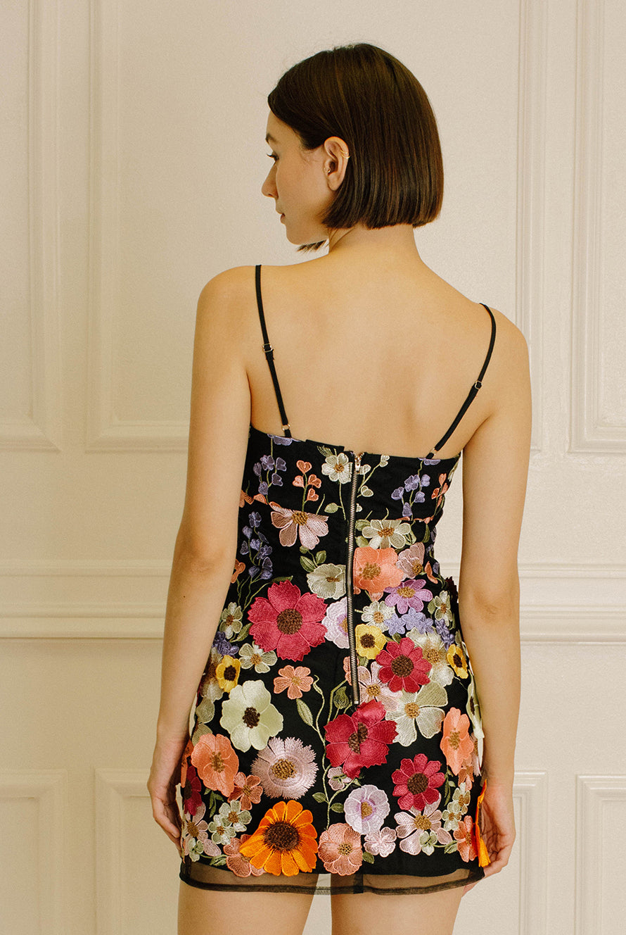 Back view of a woman wearing a black dress with colorful floral embroidery against a neutral wall.