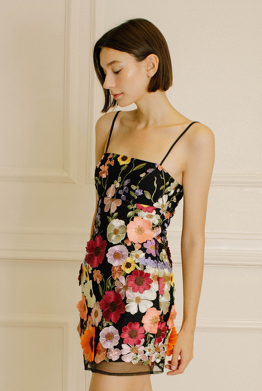 Side view of a woman wearing a black dress with colorful floral embroidery against a neutral wall.