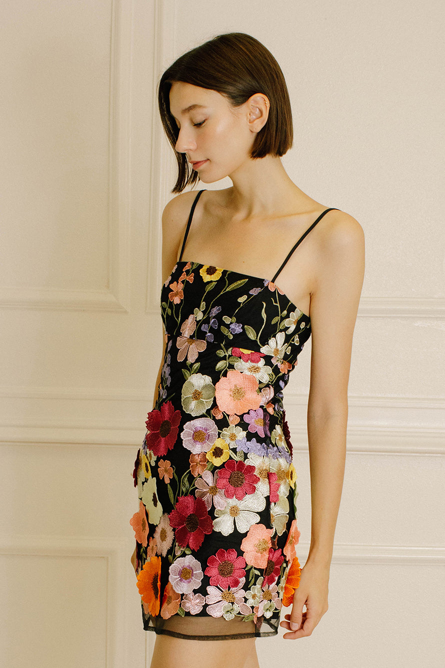 Side view of a woman wearing a black dress with colorful floral embroidery against a neutral wall.