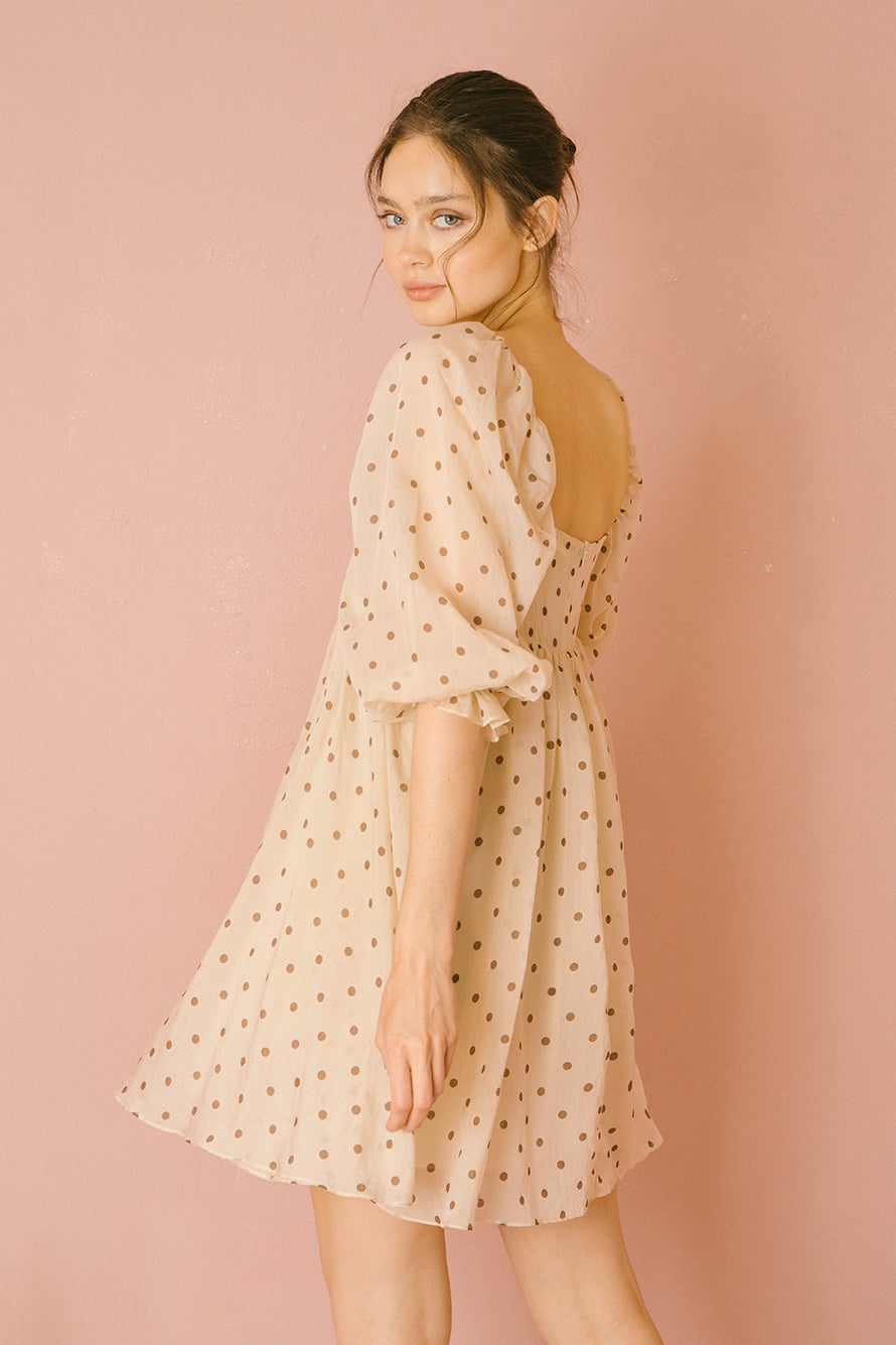 Back view of a woman wearing a beige polka dot dress against a pink background