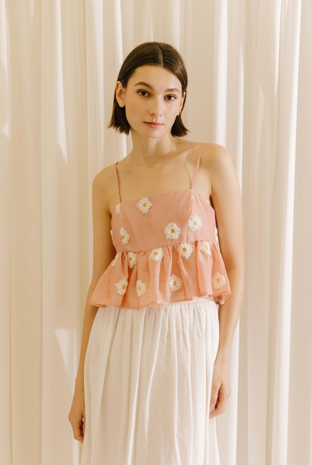 Woman wearing a pink floral top and white pants against a beige curtain background