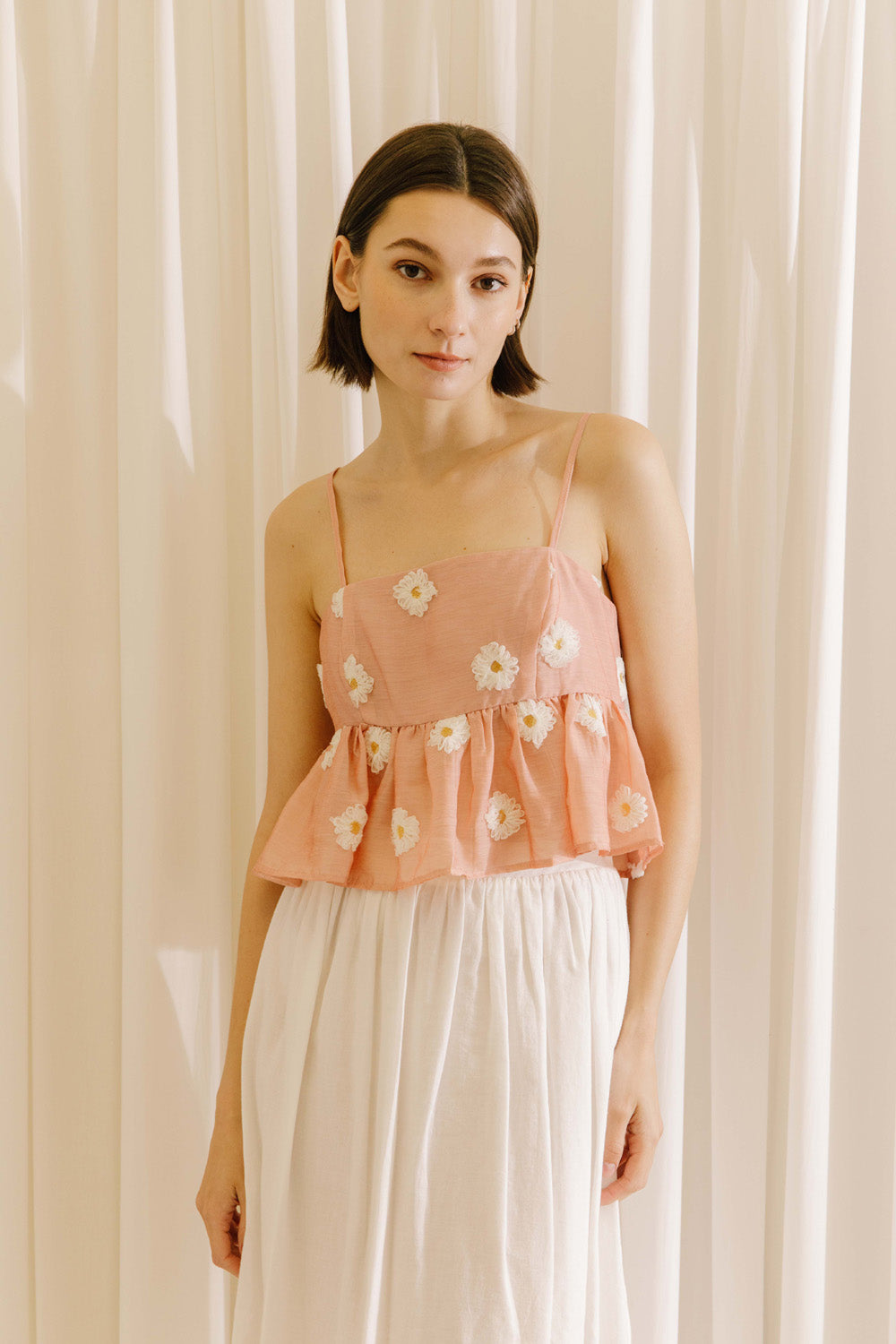 Woman wearing a pink floral top and white pants against a beige curtain background
