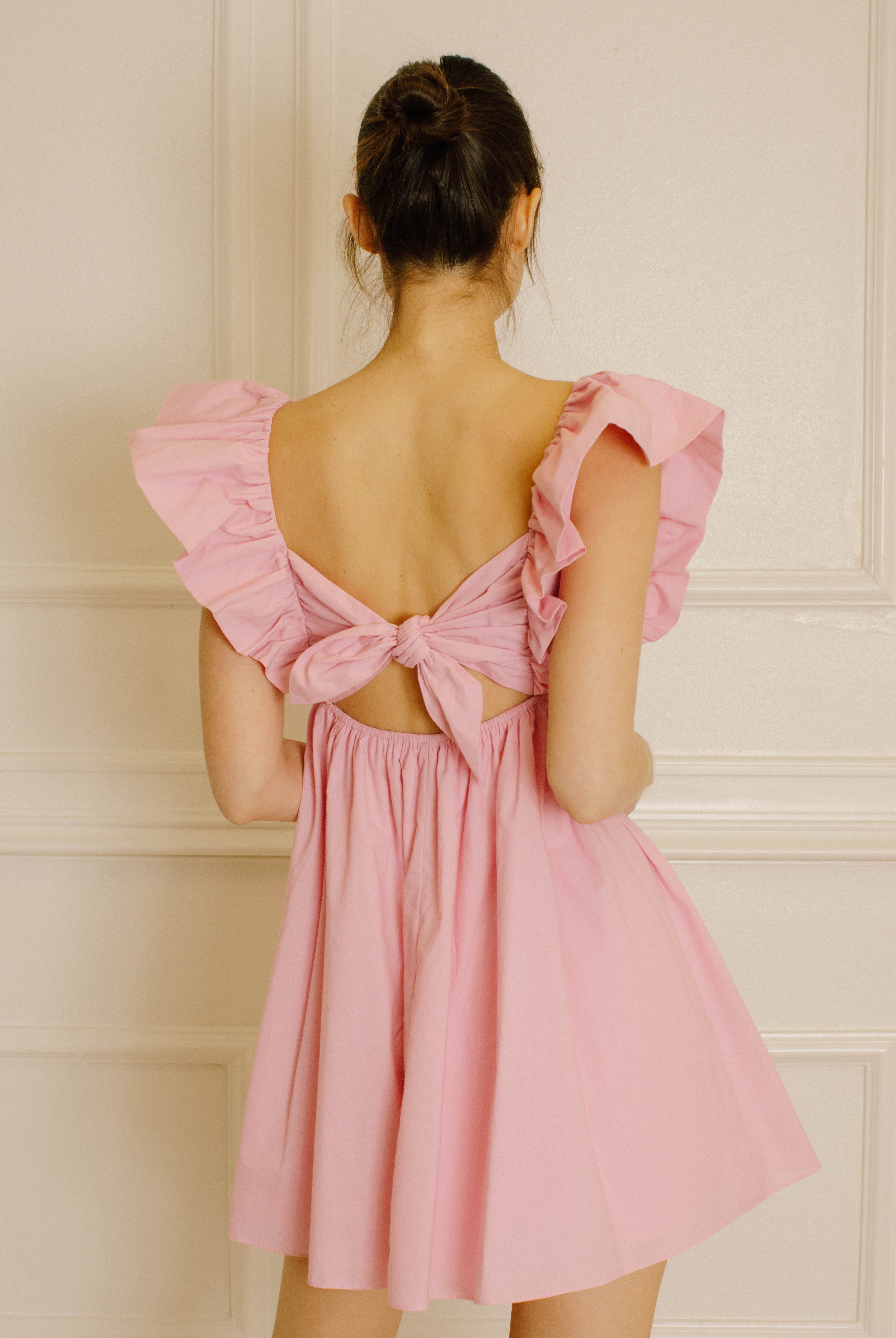 Back view of a woman wearing a pink dress with ruffled sleeves against a beige wall.