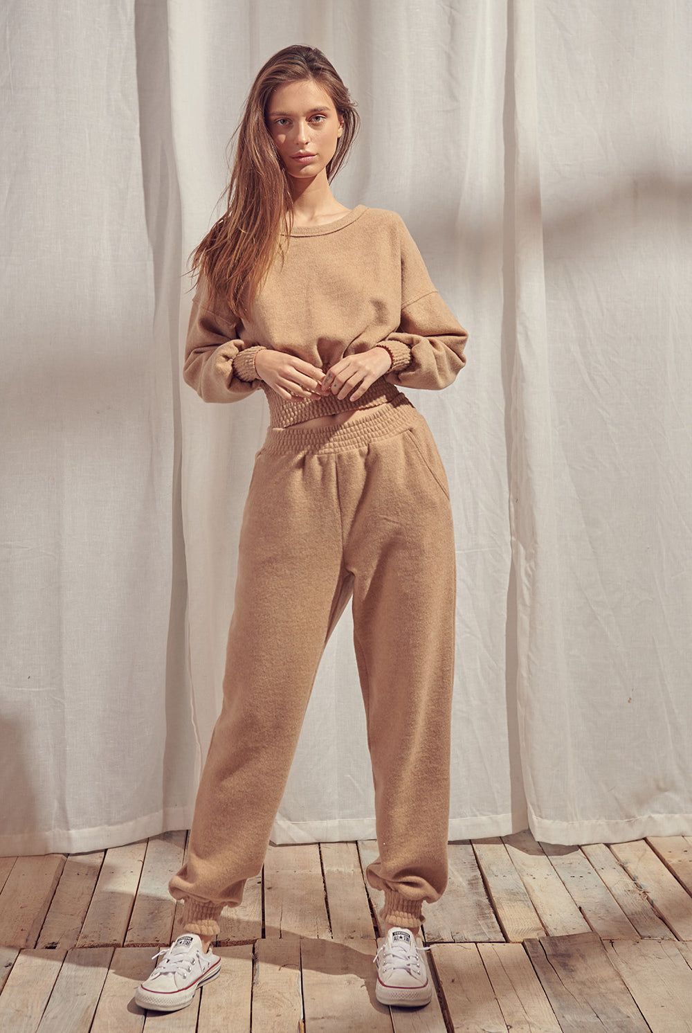 Woman wearing a beige sweater and pants standing against a white curtain.