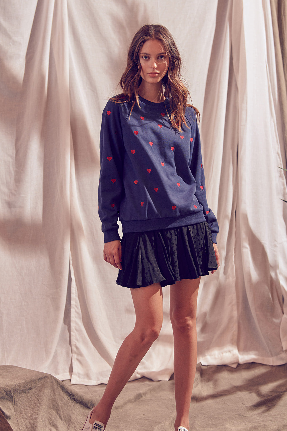 A woman standing, wearing a navy heart print pullover sweatshirt and a black skirt.