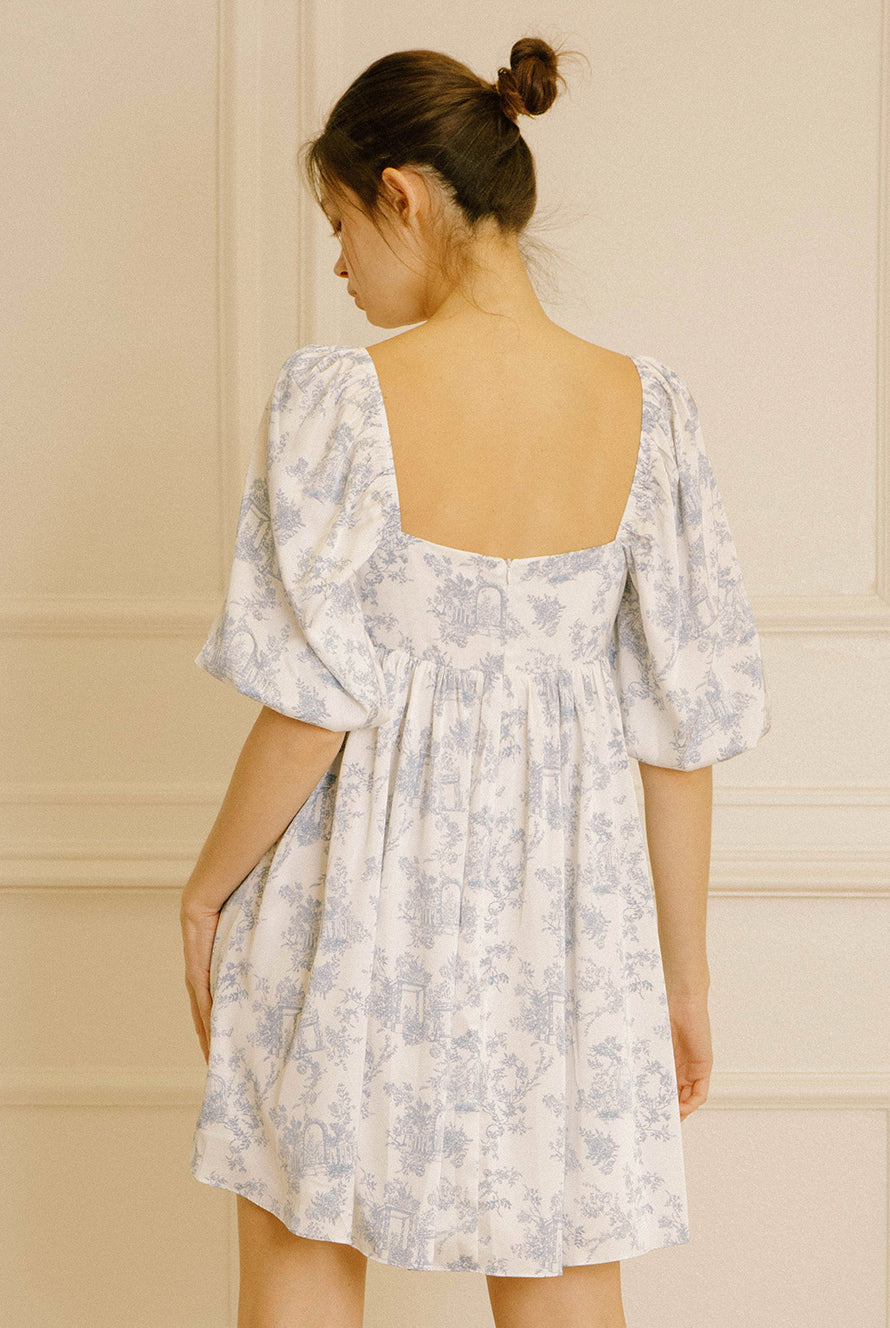 Back view of a woman wearing a white dress with a blue garden print and an invisible back zipper against a neutral background