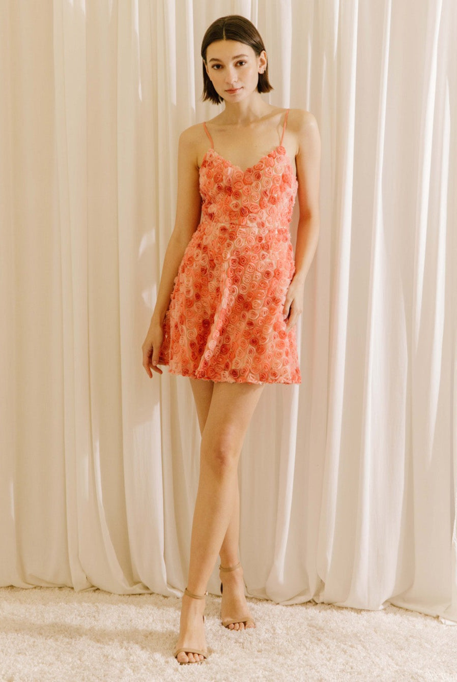 A woman standing in a white background wearing a vibrant coral 3D rose A-line mini dress with spaghetti straps and a cinched waist.