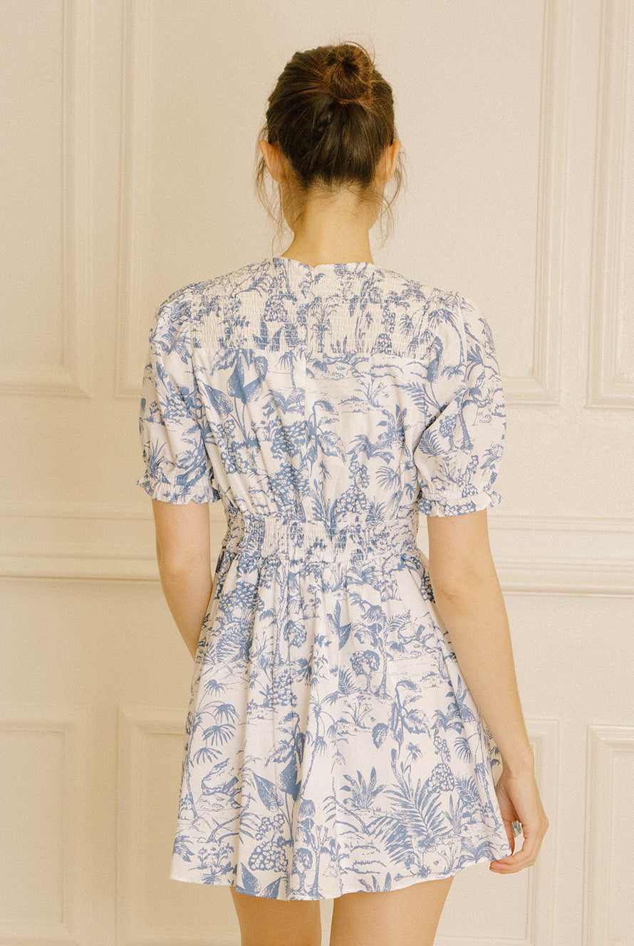 Back view of a woman wearing a blue and white tropical print mini dress against a beige wall.