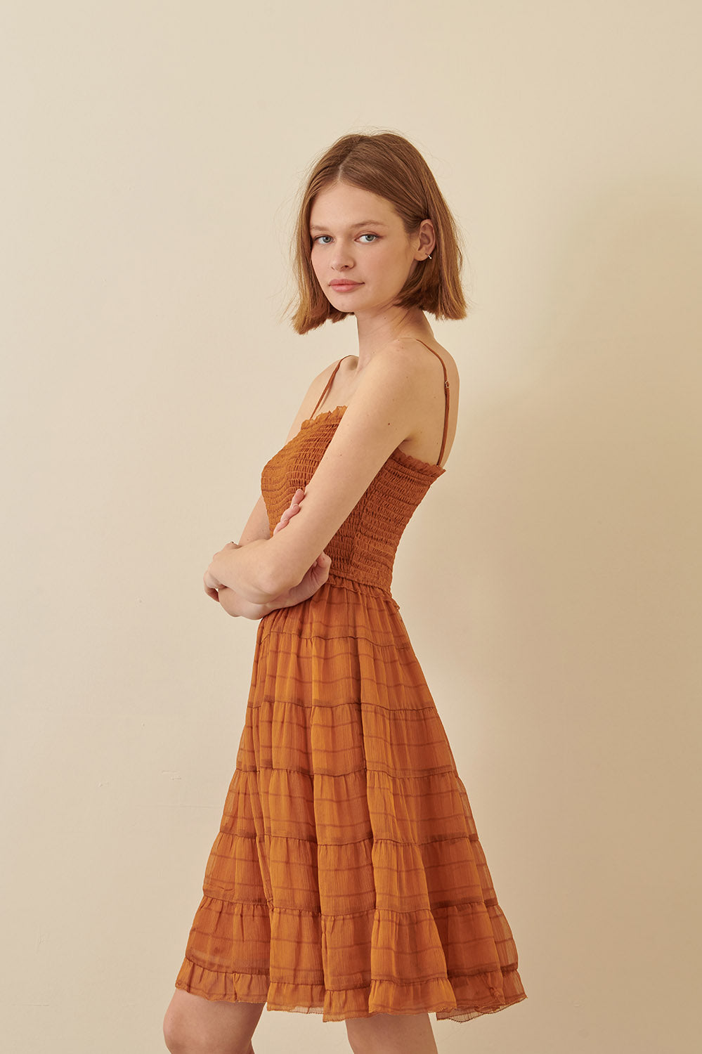Side view of a woman wearing a rust, ruffled cami dress with spaghetti straps and a straight neckline.