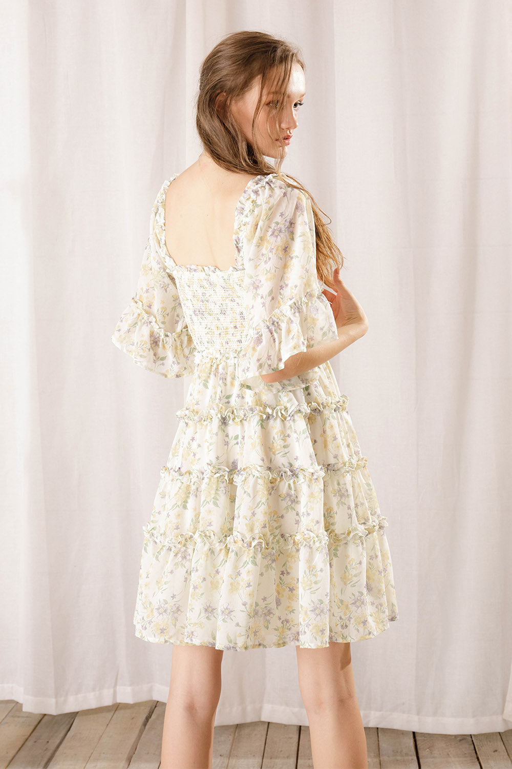 Back view of a woman wearing an ivory floral dress against a white curtain background