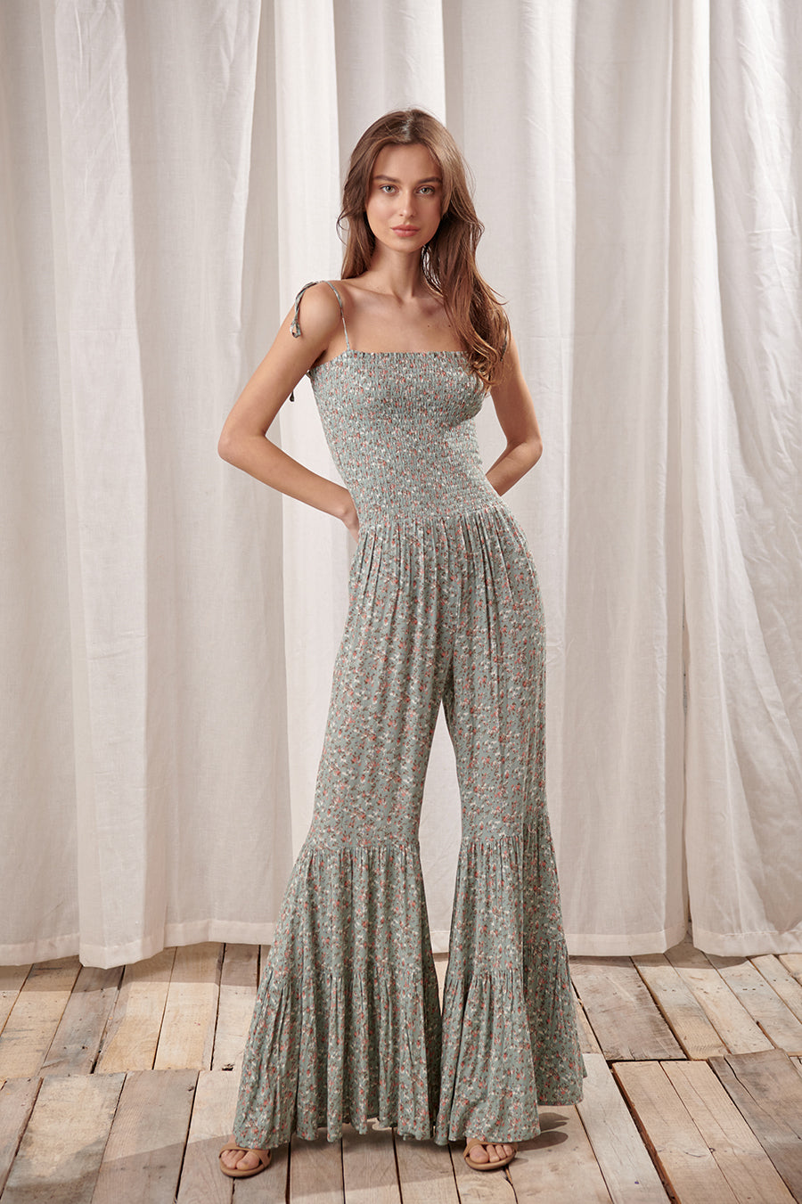 Woman wearing a green jumpsuit with a floral pattern standing in front of white curtains.
