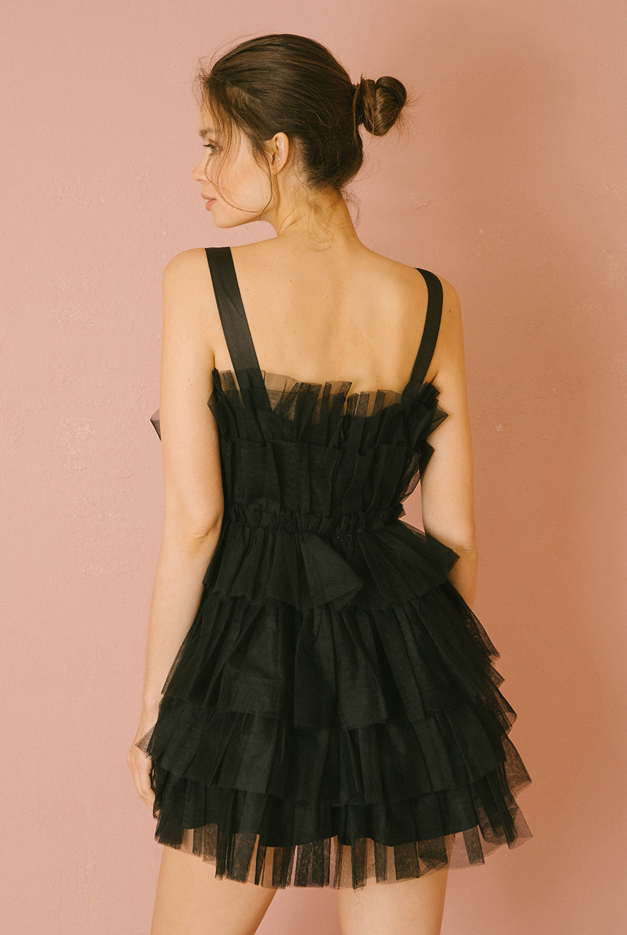 Back of a woman wearing a black tulle tiered dress against a pink background