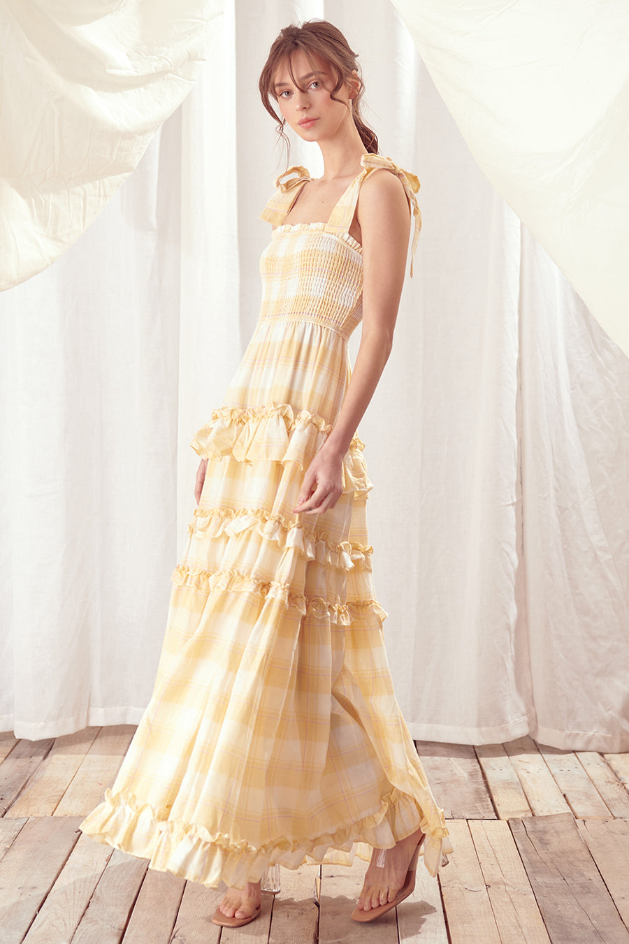 Side view of a woman wearing a yellow checkered dress standing against a white curtain.