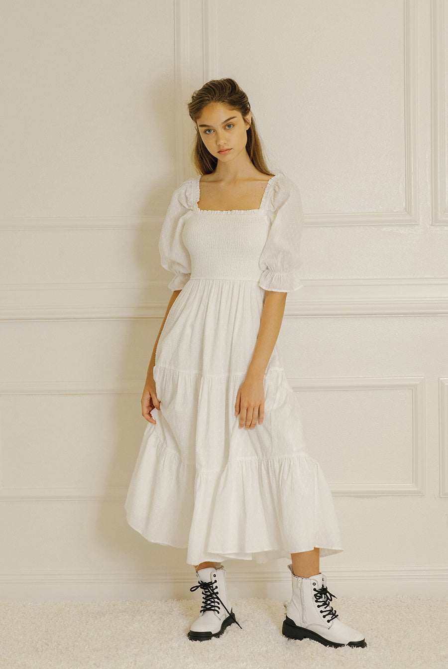 Woman wearing a white dress with puffed sleeves against a light-colored paneled wall.