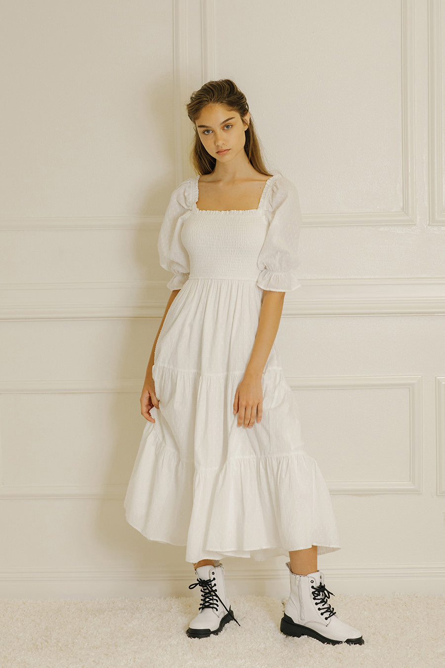 Woman wearing a white dress with puffed sleeves against a light-colored paneled wall.
