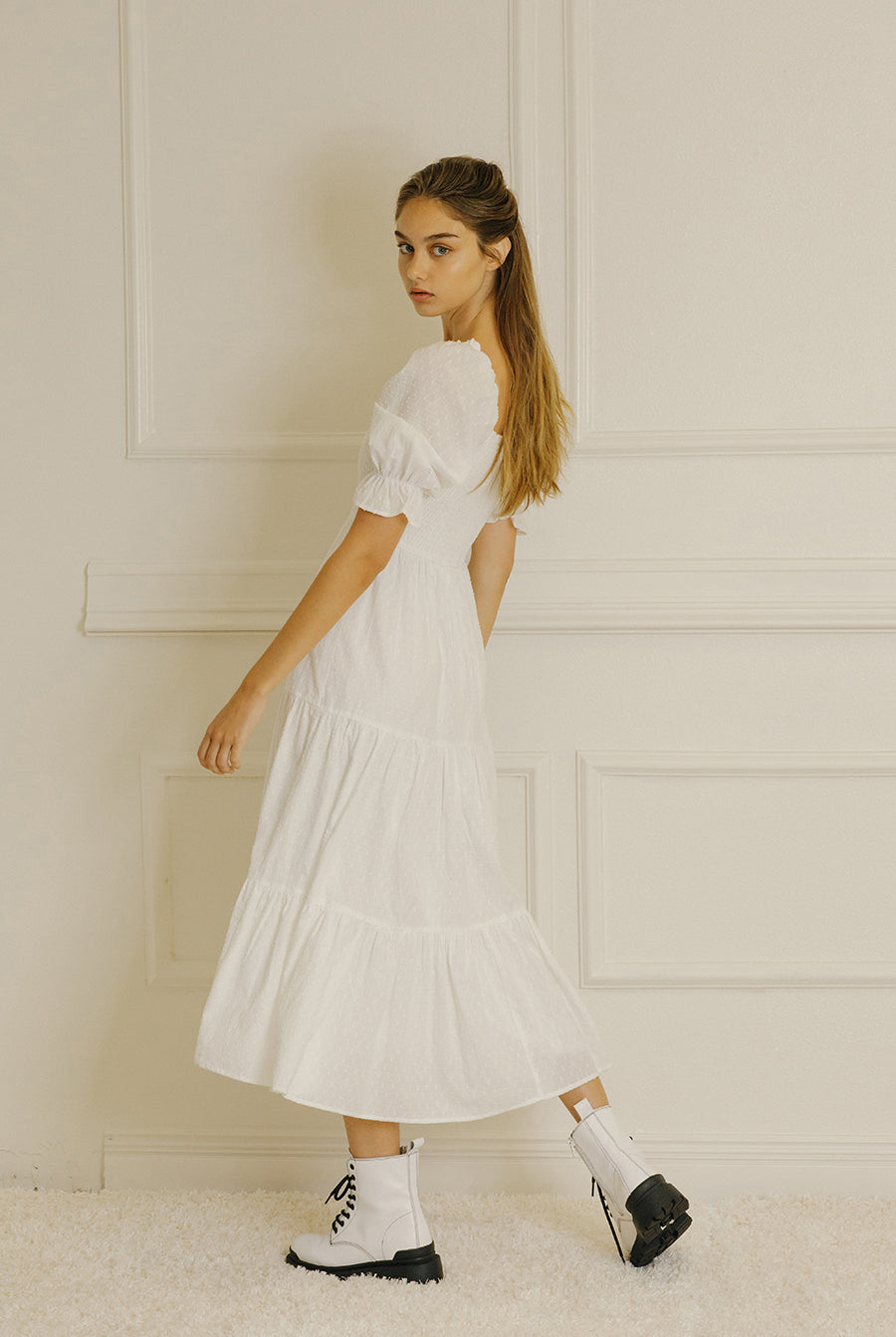 Side view of a woman wearing a white dress with puffed sleeves against a light-colored paneled wall.