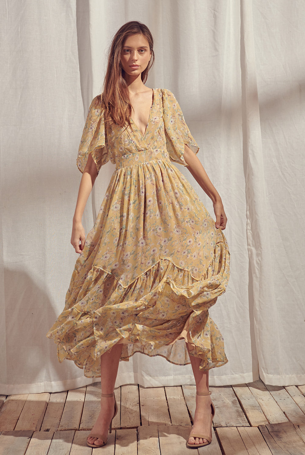 Woman wearing a yellow floral midi dress with beige high-heels standing in front of white curtains.
