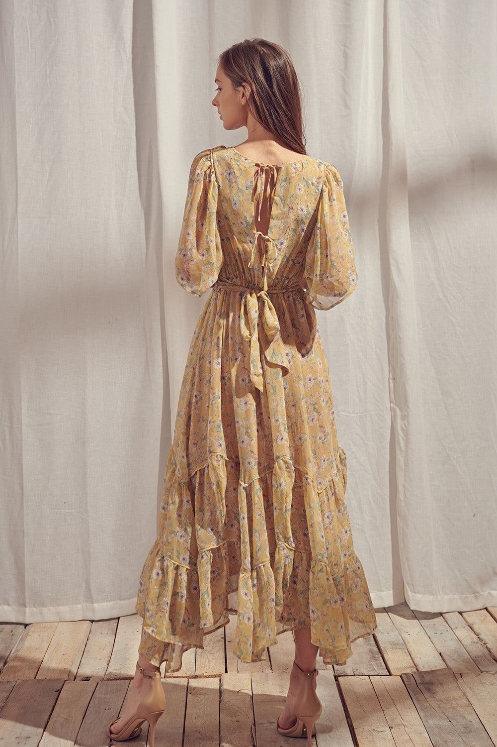Back view of a woman wearing a yellow floral midi dress with an open tie-back detail standing in front of white curtains.
