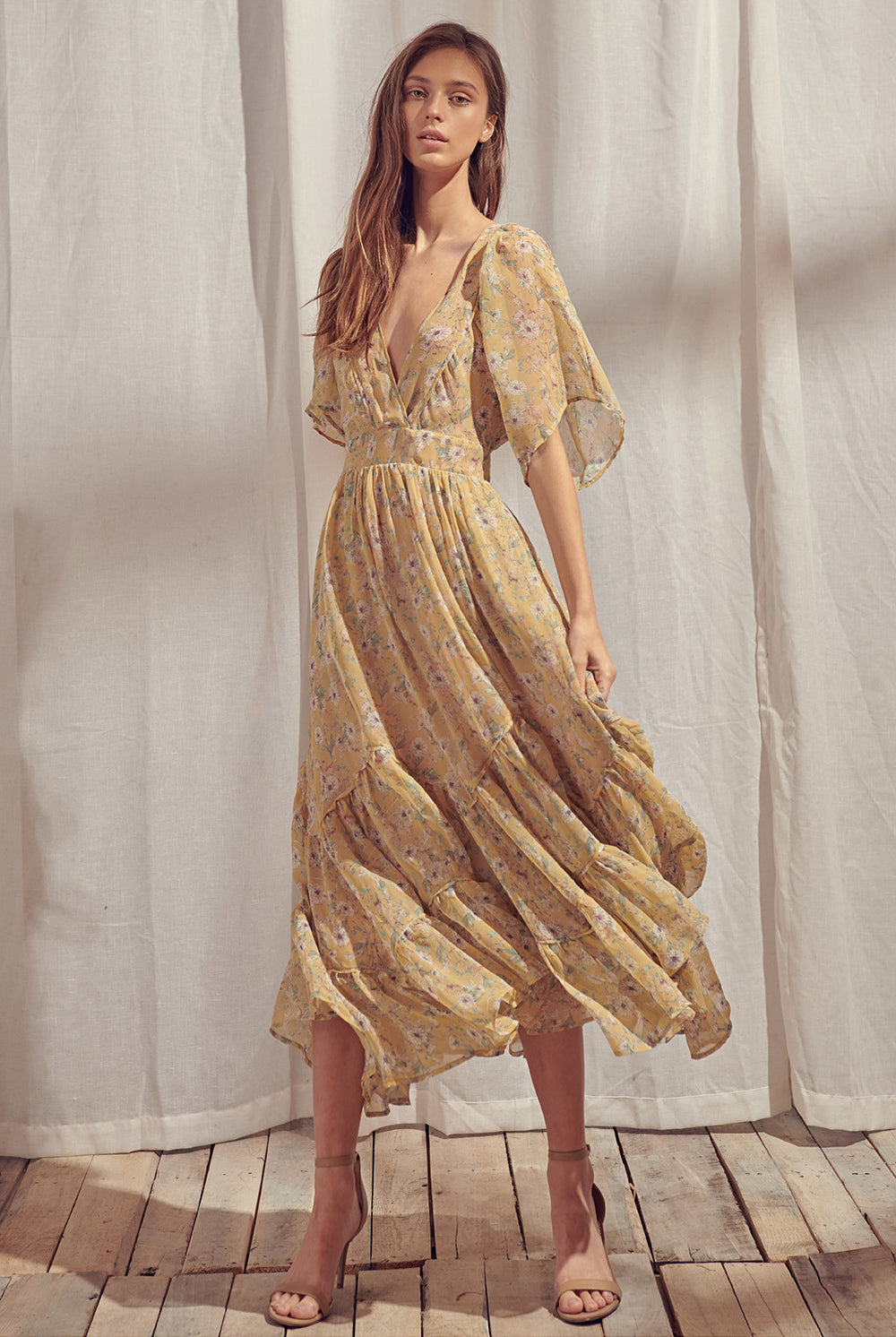 Woman wearing a yellow floral midi dress standing in front of white curtains.