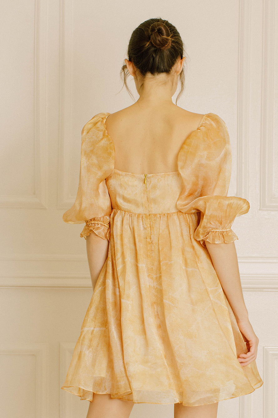 Back view of a woman wearing a yellow dress with puff sleeves against a neutral wall.